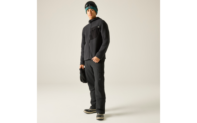 Stehende Person mit schwarzer Fleecejacke, schwarzer Hose und dunklen Stiefeln mit hellen Sohlen; hält eine schwarze Mütze; trägt eine türkise Mütze und eine Skibrille auf der Stirn; heller einfarbiger Hintergrund