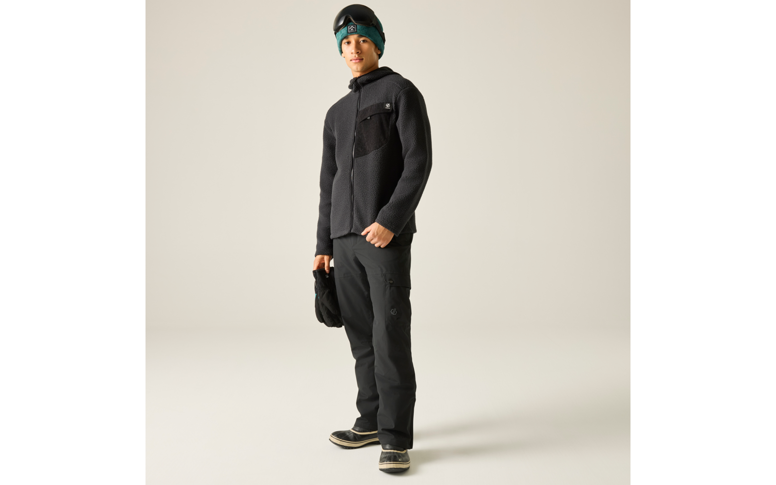 Stehende Person mit schwarzer Fleecejacke, schwarzer Hose und dunklen Stiefeln mit hellen Sohlen; hält eine schwarze Mütze; trägt eine türkise Mütze und eine Skibrille auf der Stirn; heller einfarbiger Hintergrund