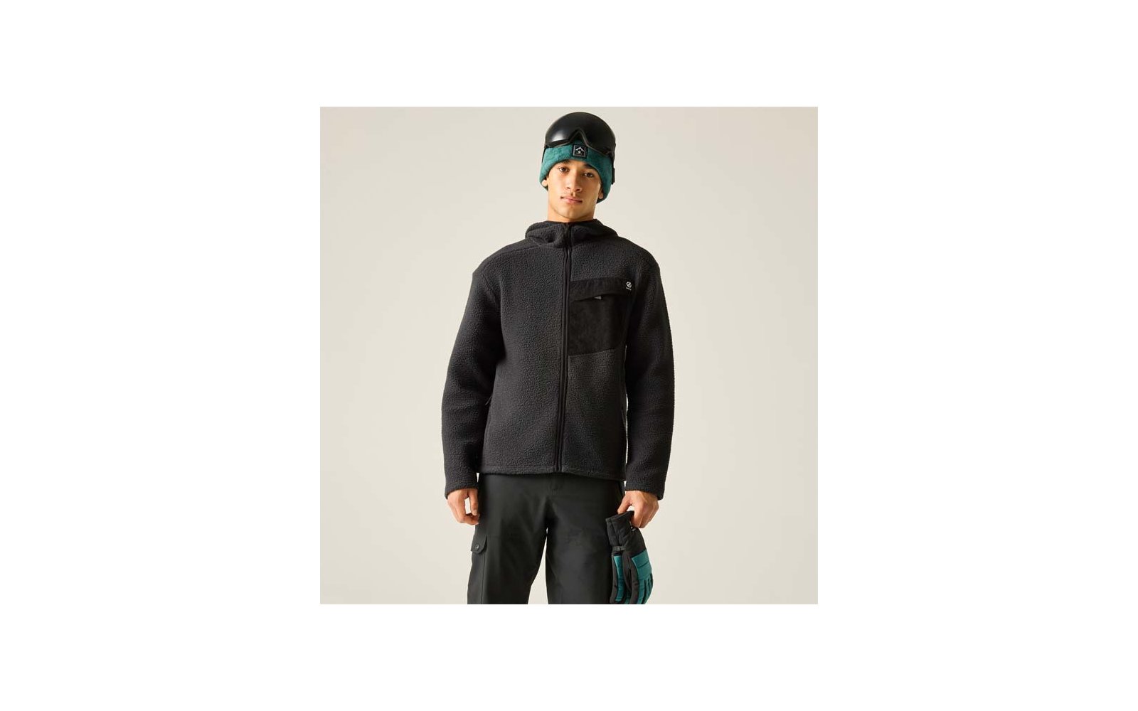 Person vor hellem Hintergrund, trägt schwarze Fleecejacke mit Brusttasche und Reißverschluss, schwarze Hose, türkise Mütze unter schwarzem Helm und hält türkise-schwarze Handschuhe