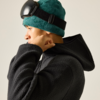 Person im Profil mit grünem Fleece-Beanie und hochgeschobener schwarzer Skibrille, schwarzer Fleece-Hoodie, Hand am Jackenkragen, heller einfarbiger Hintergrund