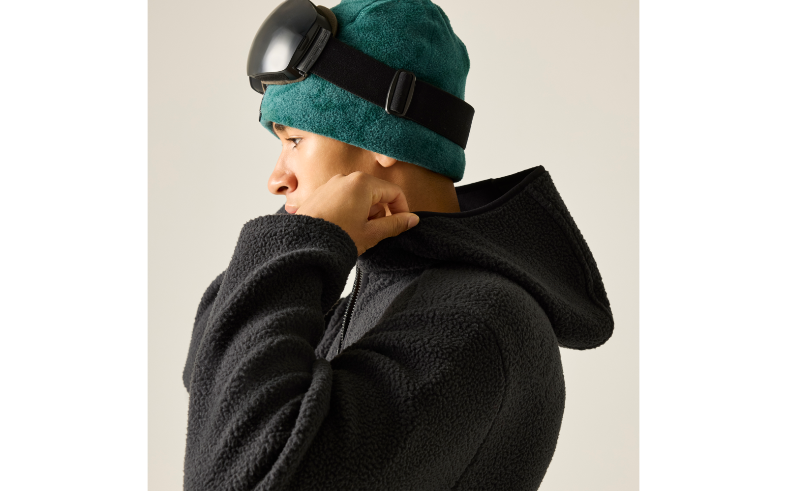 Person im Profil mit grünem Fleece-Beanie und hochgeschobener schwarzer Skibrille, schwarzer Fleece-Hoodie, Hand am Jackenkragen, heller einfarbiger Hintergrund