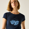 Buste d'une femme aux cheveux blonds courts et boucles d'oreilles, portant un t-shirt bleu foncé avec un motif bleu clair de montagnes et de soleil