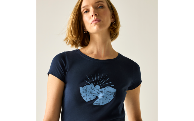 Buste d'une femme aux cheveux blonds courts et boucles d'oreilles, portant un t-shirt bleu foncé avec un motif bleu clair de montagnes et de soleil