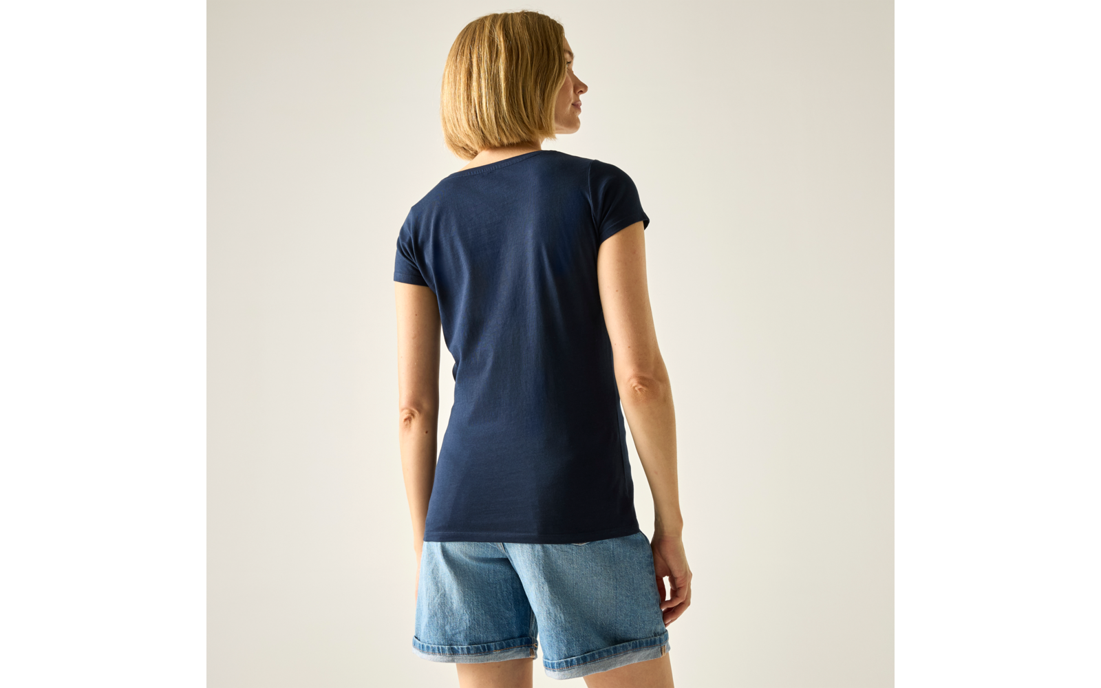 Personne de dos, porte un t-shirt bleu marine à manches courtes, cheveux blonds courts et short en jean, fond uni clair