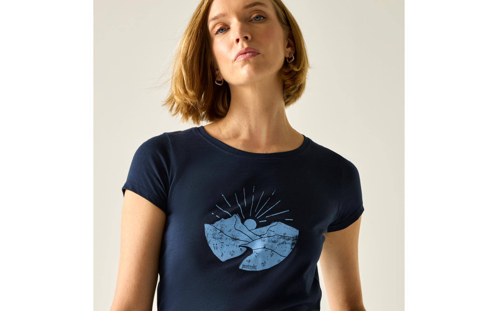 Buste d'une femme aux cheveux blonds courts et boucles d'oreilles, portant un t-shirt bleu foncé avec un motif bleu clair de montagnes et de soleil