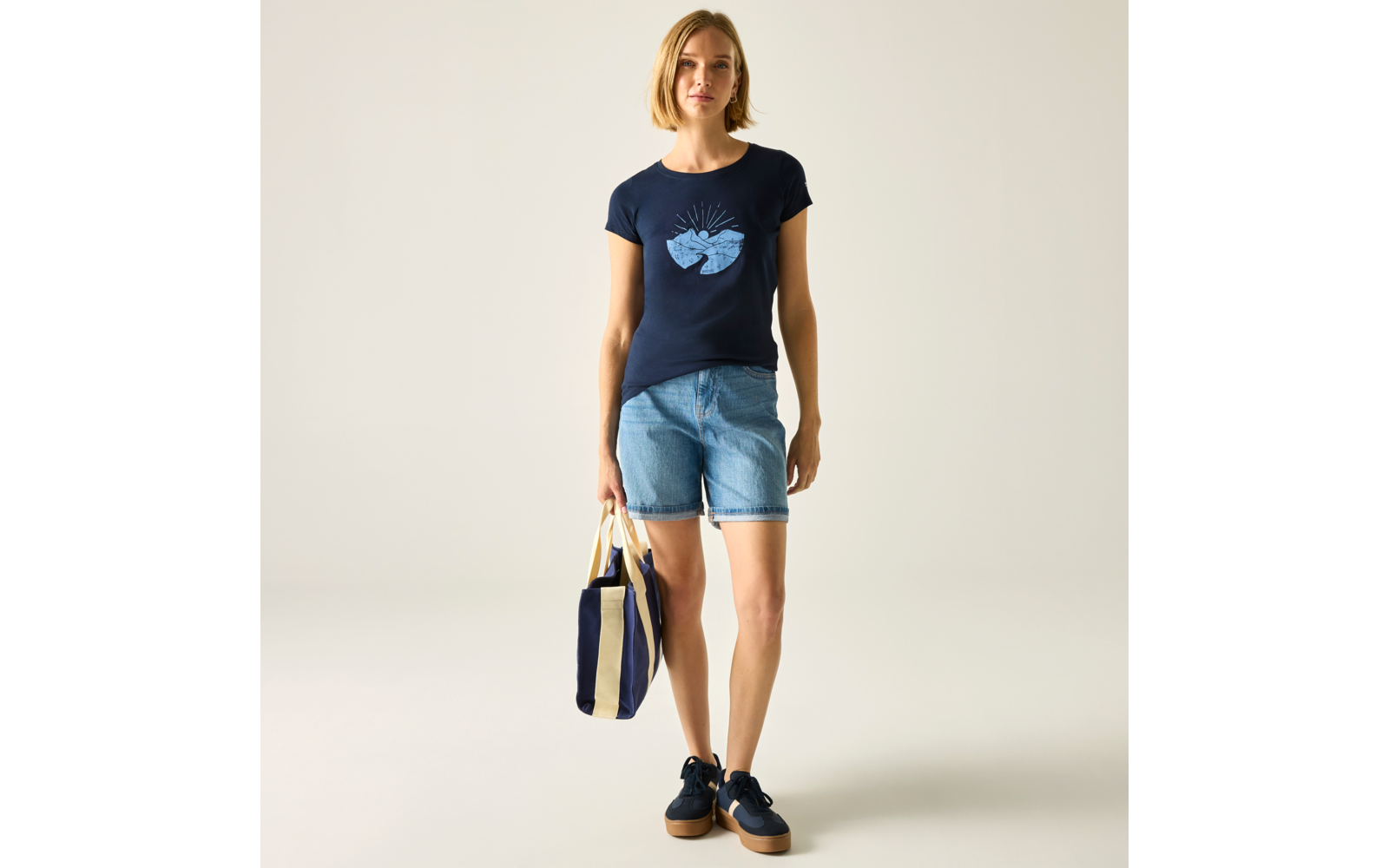 Personne debout devant un fond uni clair, porte un t‑shirt bleu marine avec un motif cœur clair, un short en jean clair, des baskets foncées et tient un sac rayé bleu et beige dans la main gauche
