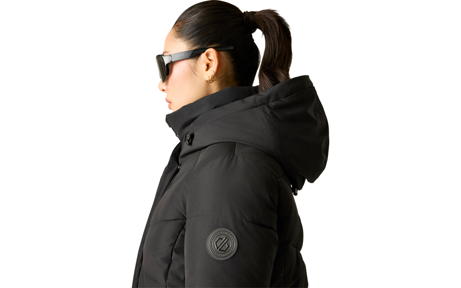 Person im Profil mit schwarzer Sonnenbrille, schwarzer gesteppter Jacke mit Kapuze, Pferdeschwanz und kleinem Ohrring