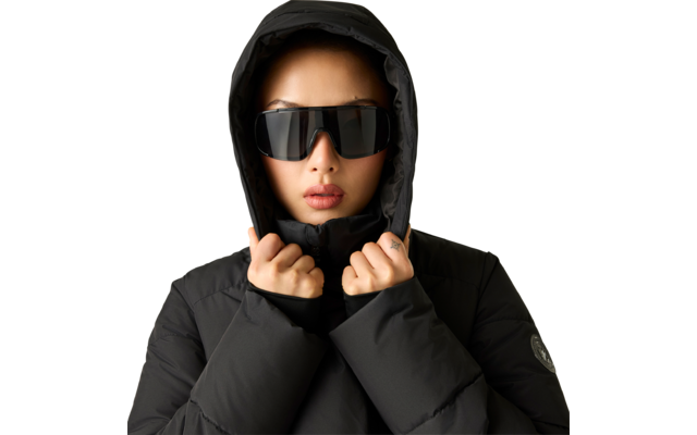 Person in schwarzer, gepolsterter Kapuzenjacke hält die Kapuze mit beiden Händen und trägt große schwarze Sonnenbrille. Neutraler heller Hintergrund