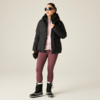 Person mit Sonnenbrille, schwarzer Steppjacke, rosa Kapuzenpullover, bordeauxfarbenen Leggings und schwarzen Winterstiefeln, hält Handschuhe, steht vor hellem Hintergrund