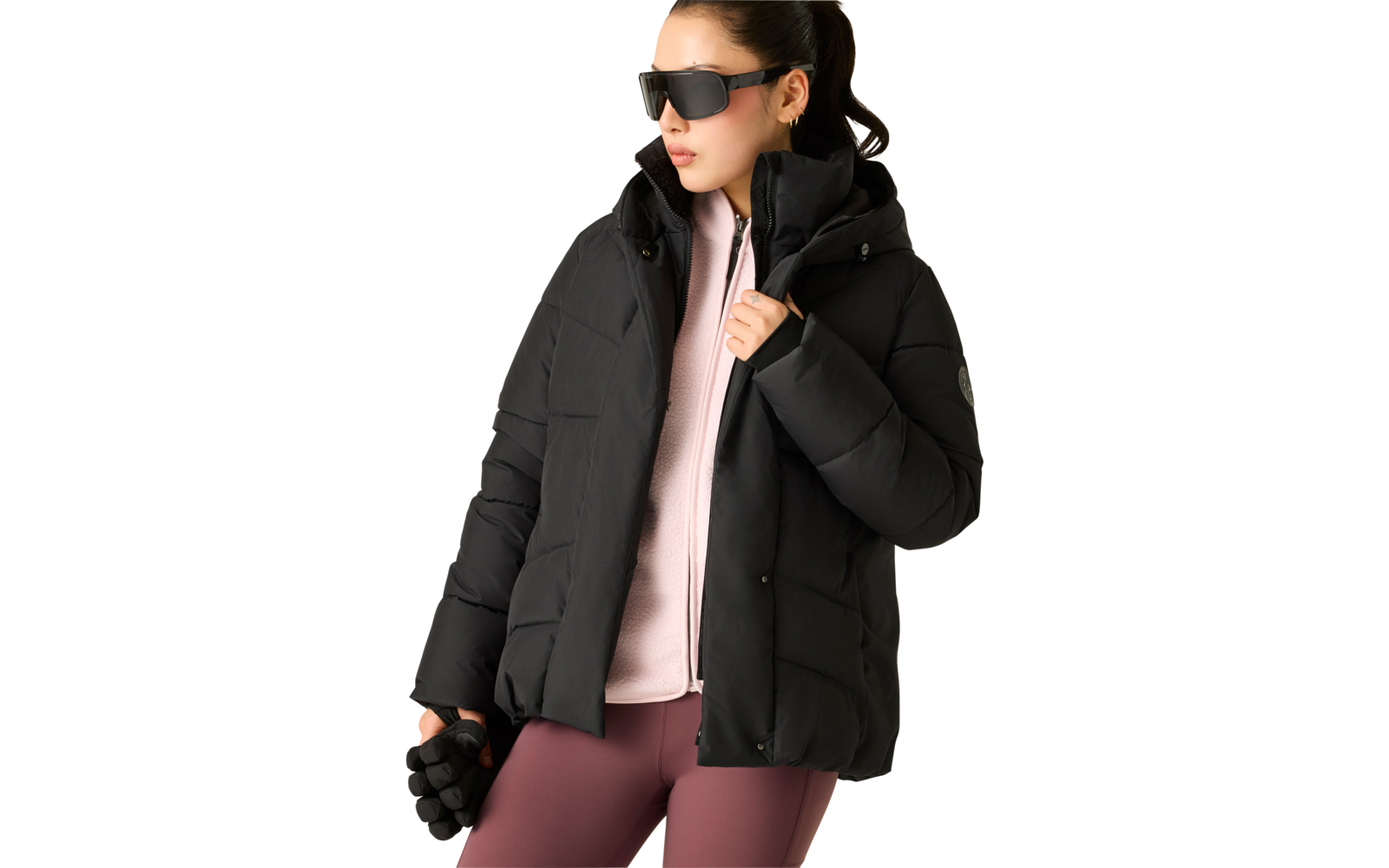Person mit zurückgebundenem Haar, trägt schwarze Steppjacke mit Kapuze, rosa Zip-Sweater, bordeauxfarbene Leggings, große schwarze Sonnenbrille, hält schwarze Handschuhe in der linken Hand