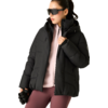 Person mit zurückgebundenem Haar, trägt schwarze Steppjacke mit Kapuze, rosa Zip-Sweater, bordeauxfarbene Leggings, große schwarze Sonnenbrille, hält schwarze Handschuhe in der linken Hand