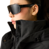 Nahaufnahme einer Person mit zurückgebundenen Haaren, schwarzer Sonnenbrille, drei kleinen Ohrringen und schwarzer Steppjacke mit hohem Kragen