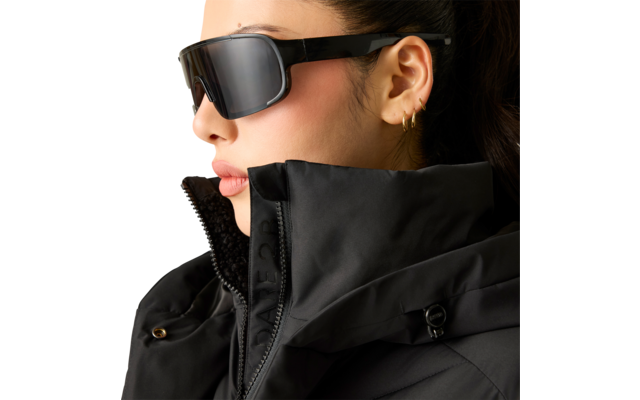 Nahaufnahme einer Person mit zurückgebundenen Haaren, schwarzer Sonnenbrille, drei kleinen Ohrringen und schwarzer Steppjacke mit hohem Kragen