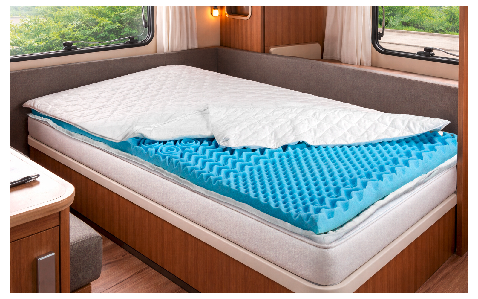 Ein Einzelbett in einem Wohnmobil mit weißer gesteppter Auflage teilweise zurückgefaltet, blauem Noppen-Schaumstofftopper, Holzverkleidung, zwei Fenstern mit Gardinen und Blick auf grüne Pflanzen