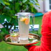 Glas mit Zitronenwasser und Glas mit Eiswürfeln auf goldenem Tablett, Person mit rotem Pullover im Hintergrund