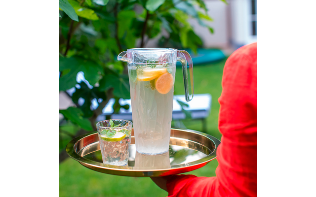 Glas mit Zitronenwasser und Glas mit Eiswürfeln auf goldenem Tablett, Person mit rotem Pullover im Hintergrund