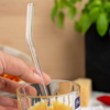 Hand hält gebogenen Glasstrohhalm in Glas mit orangefarbener Flüssigkeit; blaues Etikett am Glas; unscharfe Topfpflanze und Gegenstände im Hintergrund
