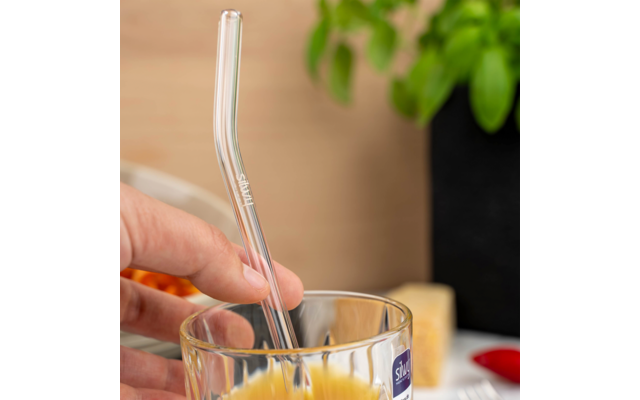 Hand hält gebogenen Glasstrohhalm in Glas mit orangefarbener Flüssigkeit; blaues Etikett am Glas; unscharfe Topfpflanze und Gegenstände im Hintergrund