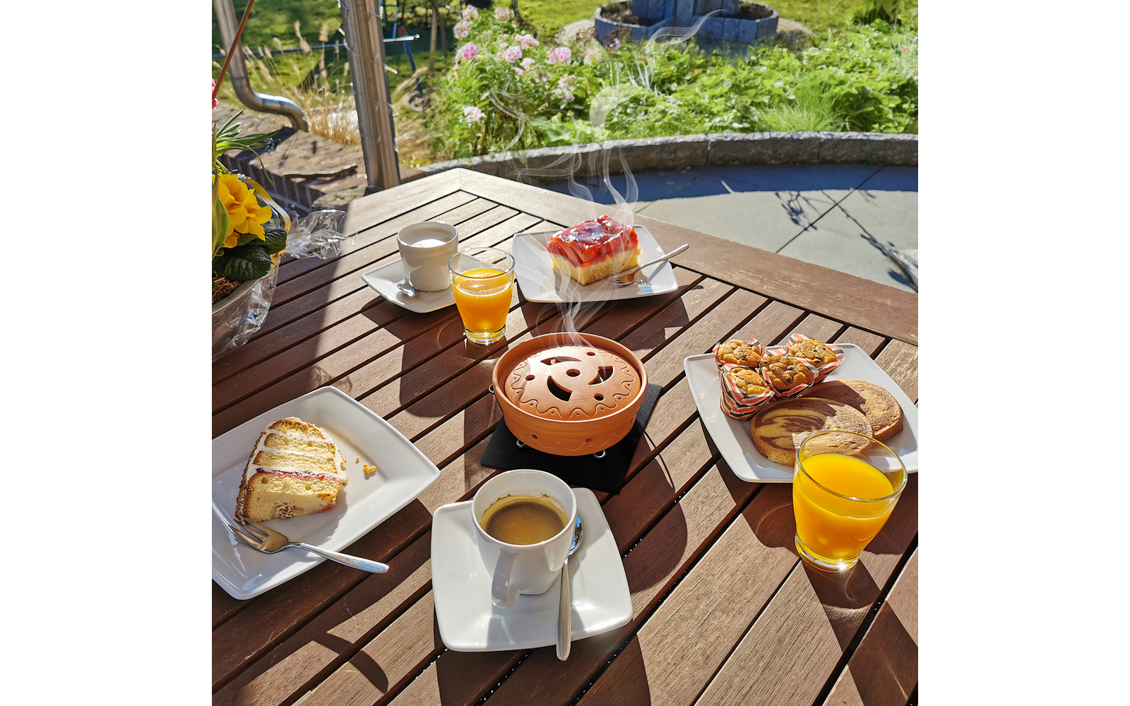 Holztisch mit mehreren Tellern, einer Kaffeetasse, einem Glas Orangensaft und einer Teekanne im Freien