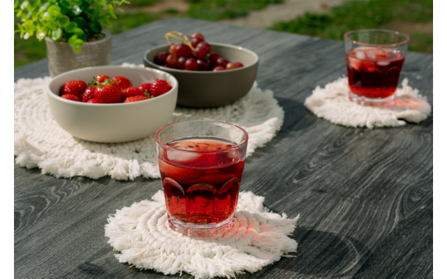 Mesa de madera oscura con tres vasos con bebida roja y cubitos de hielo sobre posavasos redondos blancos con flecos; dos cuencos con fresas y cerezas y una pequeña planta en maceta al fondo