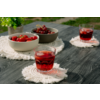 Mesa de madera oscura con tres vasos con bebida roja y cubitos de hielo sobre posavasos redondos blancos con flecos; dos cuencos con fresas y cerezas y una pequeña planta en maceta al fondo