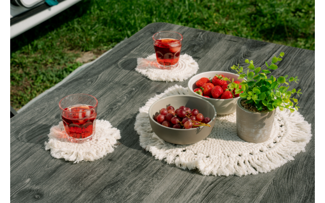 Mesa de madera gris al aire libre con dos vasos con bebida roja sobre posavasos macramé blancos, dos cuencos con uvas y fresas, una pequeña maceta con planta verde y un mantelito blanco de ganchillo; césped al fondo