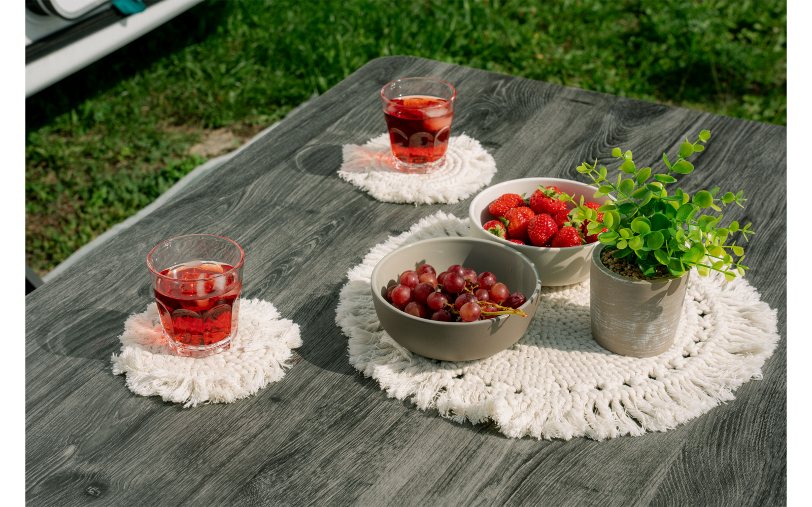Mesa de madera gris al aire libre con dos vasos con bebida roja sobre posavasos macramé blancos, dos cuencos con uvas y fresas, una pequeña maceta con planta verde y un mantelito blanco de ganchillo; césped al fondo