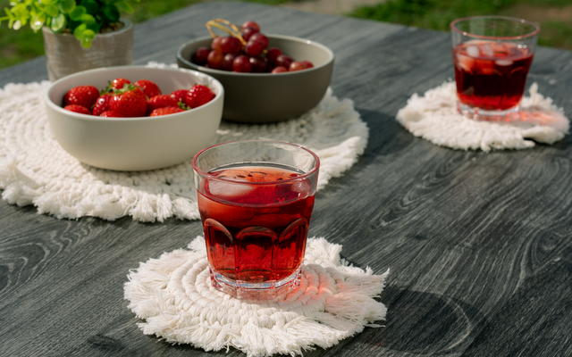 Dunkler Holztisch mit drei Gläsern mit rotem Getränk und Eiswürfeln auf weißen, runden Untersetzern mit Fransen; zwei Schalen mit Erdbeeren und Kirschen und eine kleine Topfpflanze im Hintergrund