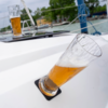 Tilted beer glass with some foam in a black drink holder on a white boat deck; in the background a second beer glass, rigging, water and a shoreline with trees