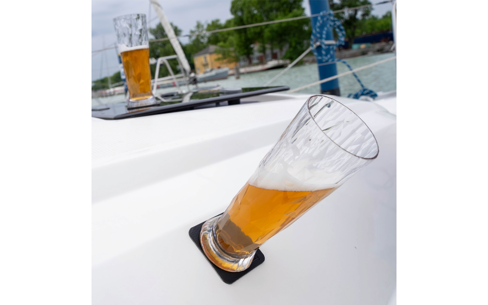 Tilted beer glass with some foam in a black drink holder on a white boat deck; in the background a second beer glass, rigging, water and a shoreline with trees