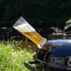 Tilted tall beer glass with pale beer and foam inserted into the vent of a black kettle grill; river and green shoreline vegetation in the background; reflections of a building and a person on the grill lid