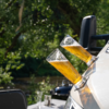 Two tilted beer glasses with foam in holders on a white boat side, blurred river and trees in the background