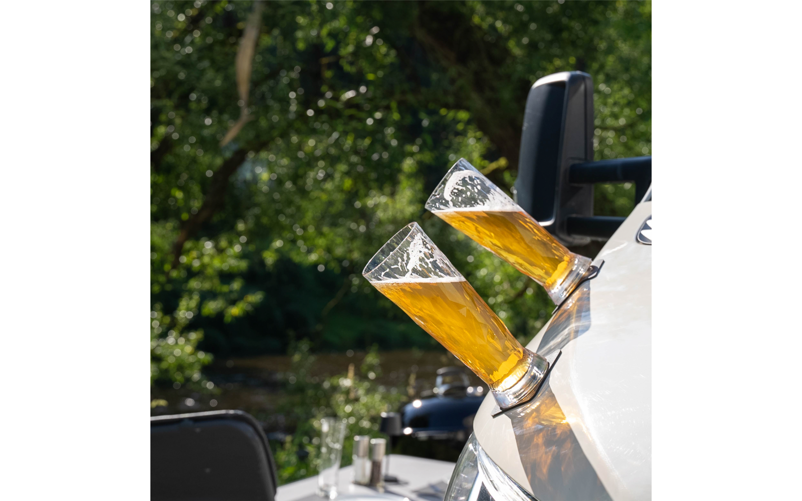 Two tilted beer glasses with foam in holders on a white boat side, blurred river and trees in the background