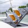 Two tall beer glasses on a tilted black boat hatch, background shows water, moored boats and trees under a cloudy sky