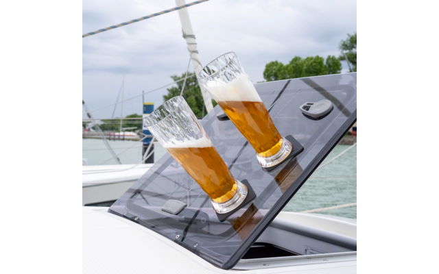 Two tall beer glasses on a tilted black boat hatch, background shows water, moored boats and trees under a cloudy sky