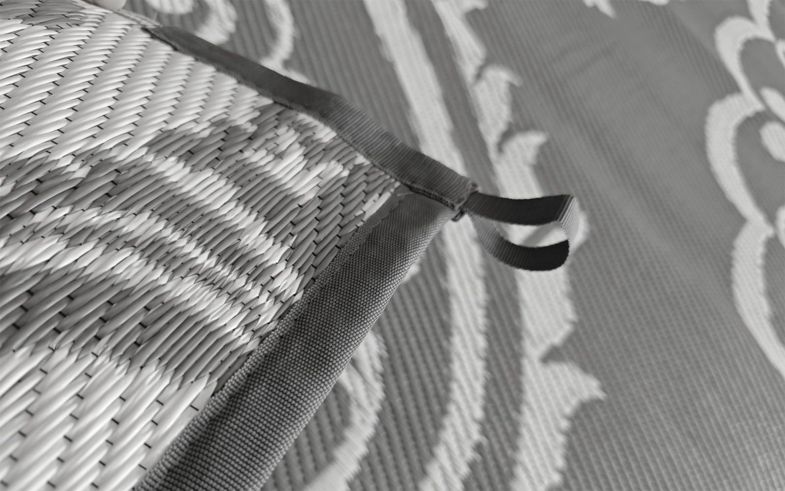 Nahaufnahme einer geflochtenen Oberfläche mit Stoffkante und kleiner Schlaufe; im Hintergrund ein gemusterter Teppich mit kreisförmigen Ornamenten