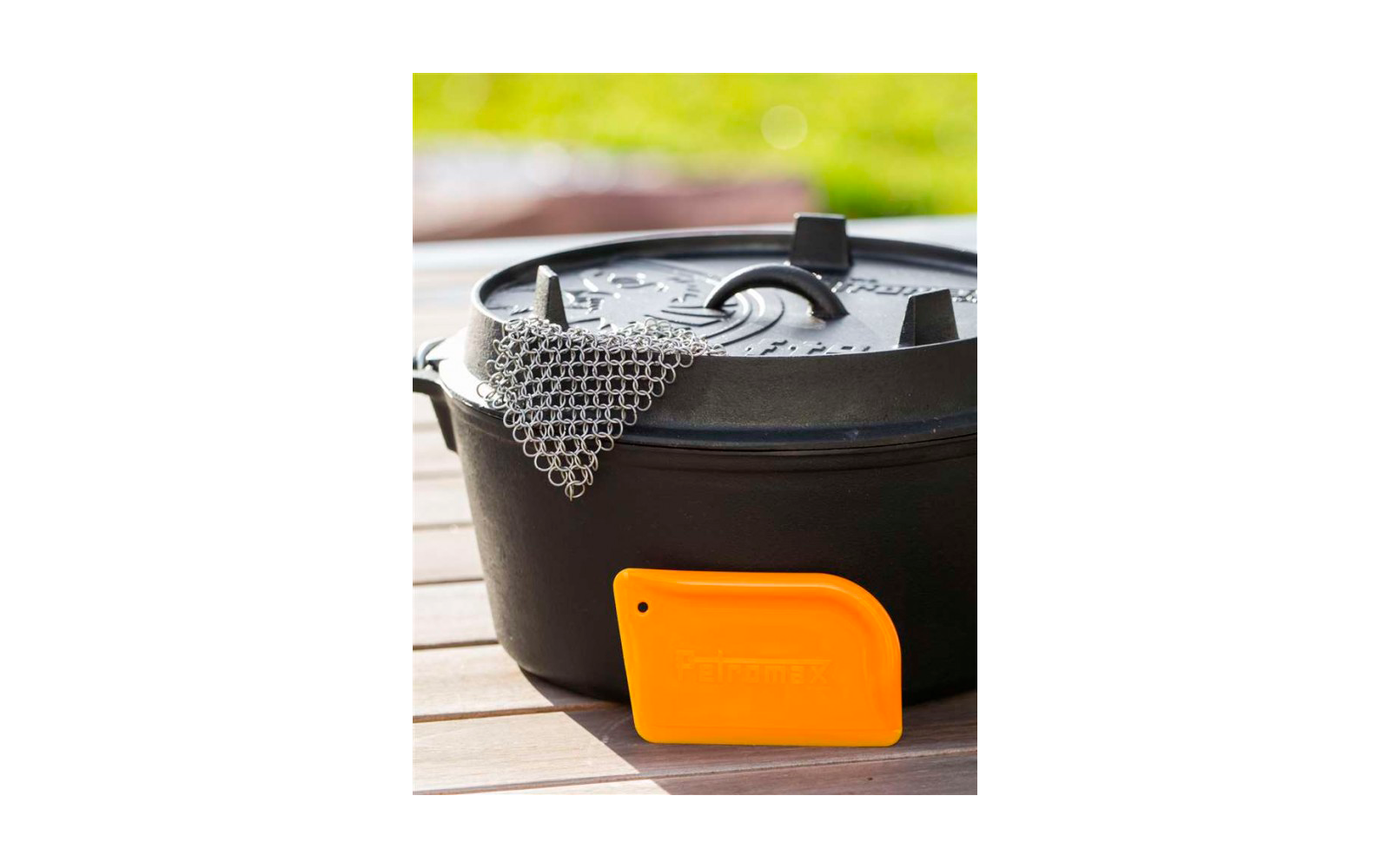Ein schwarzer Topf mit Deckel und einem grauen Topflappen liegt auf einer Holzfläche. Vor dem Topf steht ein oranger Kunststoffgegenstand