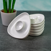 Stacked white triangular dishes with circular depressions on a dark surface, a potted green plant visible in the background left