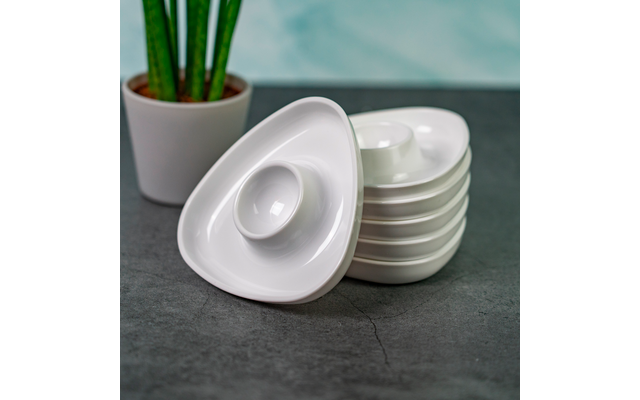 Stacked white triangular dishes with circular depressions on a dark surface, a potted green plant visible in the background left