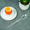 Opened egg in eggshell with visible runny yolk on a white saucer, transparent plastic spoon beside it, green succulent plant in a white pot on a gray surface