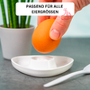 Hand holding brown egg above a white egg cup on a gray surface; white spoon to the right, potted plant blurred in the background left; white rectangle with the text 'PASSEND FÜR ALLE EIERGRÖSSEN' and a thumbs-up icon at the top