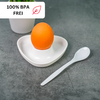 Orange egg in a white heart-shaped dish on a gray surface, a white spoon beside it; a white cup in the background and a label at top left reading "100% BPA FREI" with a leaf icon