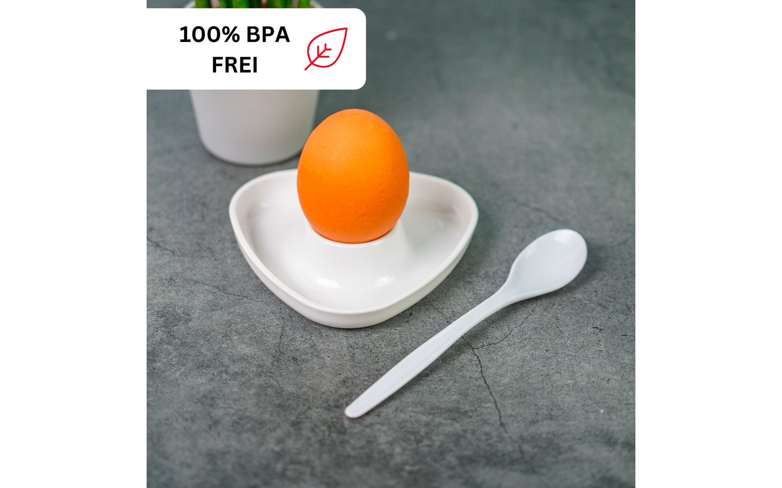 Orange egg in a white heart-shaped dish on a gray surface, a white spoon beside it; a white cup in the background and a label at top left reading "100% BPA FREI" with a leaf icon