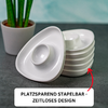 Stacked white triangular bowls with a central recess, one bowl tilted to the side; red arrow pointing at the stack; white rectangular label with black text "PLATZSPAREND STAPELBAR - ZEITLOSES DESIGN"; left a green potted plant in a light pot; gray tabletop, blurred turquoise background