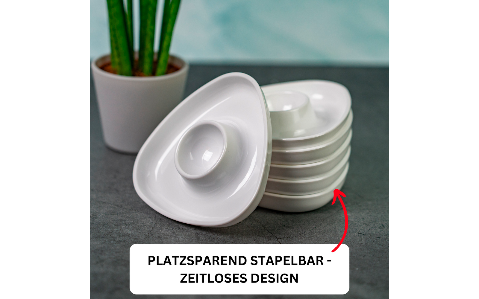 Stacked white triangular bowls with a central recess, one bowl tilted to the side; red arrow pointing at the stack; white rectangular label with black text "PLATZSPAREND STAPELBAR - ZEITLOSES DESIGN"; left a green potted plant in a light pot; gray tabletop, blurred turquoise background
