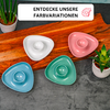 Four triangular glossy ceramic dishes with a central round indentation in white, pink, green and light blue on a wooden board on a gray concrete surface. A small potted plant top left and bottom right. Above, a white banner with black text 'ENTDECKE UNSERE FARBVARIATIONEN' and a red paint-palette icon