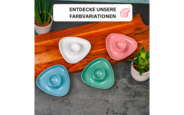 Four triangular glossy ceramic dishes with a central round indentation in white, pink, green and light blue on a wooden board on a gray concrete surface. A small potted plant top left and bottom right. Above, a white banner with black text 'ENTDECKE UNSERE FARBVARIATIONEN' and a red paint-palette icon