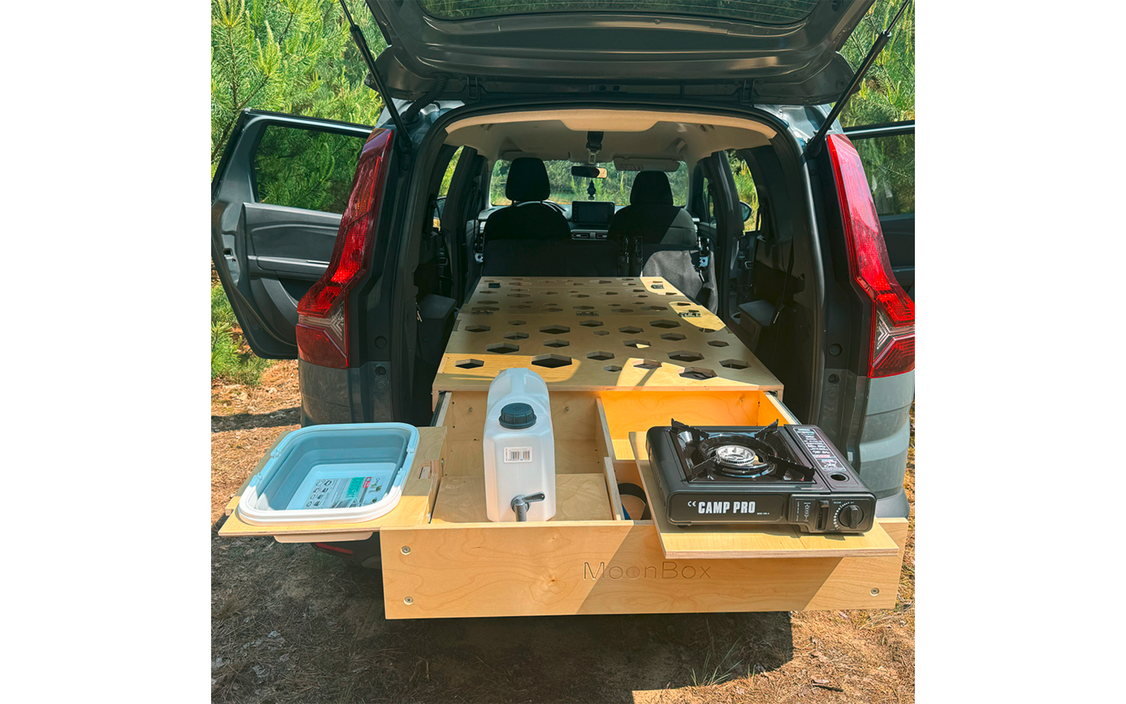 Offener Kofferraum eines Fahrzeugs mit einer herausgezogenen Holzkonstruktion mit runden Aussparungen, links eine blaue Plastikwanne, mittig ein weißer Wasserkanister, rechts ein tragbarer Gaskocher, zwei Vordersitze und Nadelbäume im Hintergrund