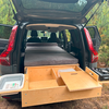 Offenes Heck eines SUVs mit grauer Matratze auf einer Plattform, herausziehbare Holzschublade mit Fächern, Plastikwanne links, Campingkocher rechts, Nadelbäume im Hintergrund