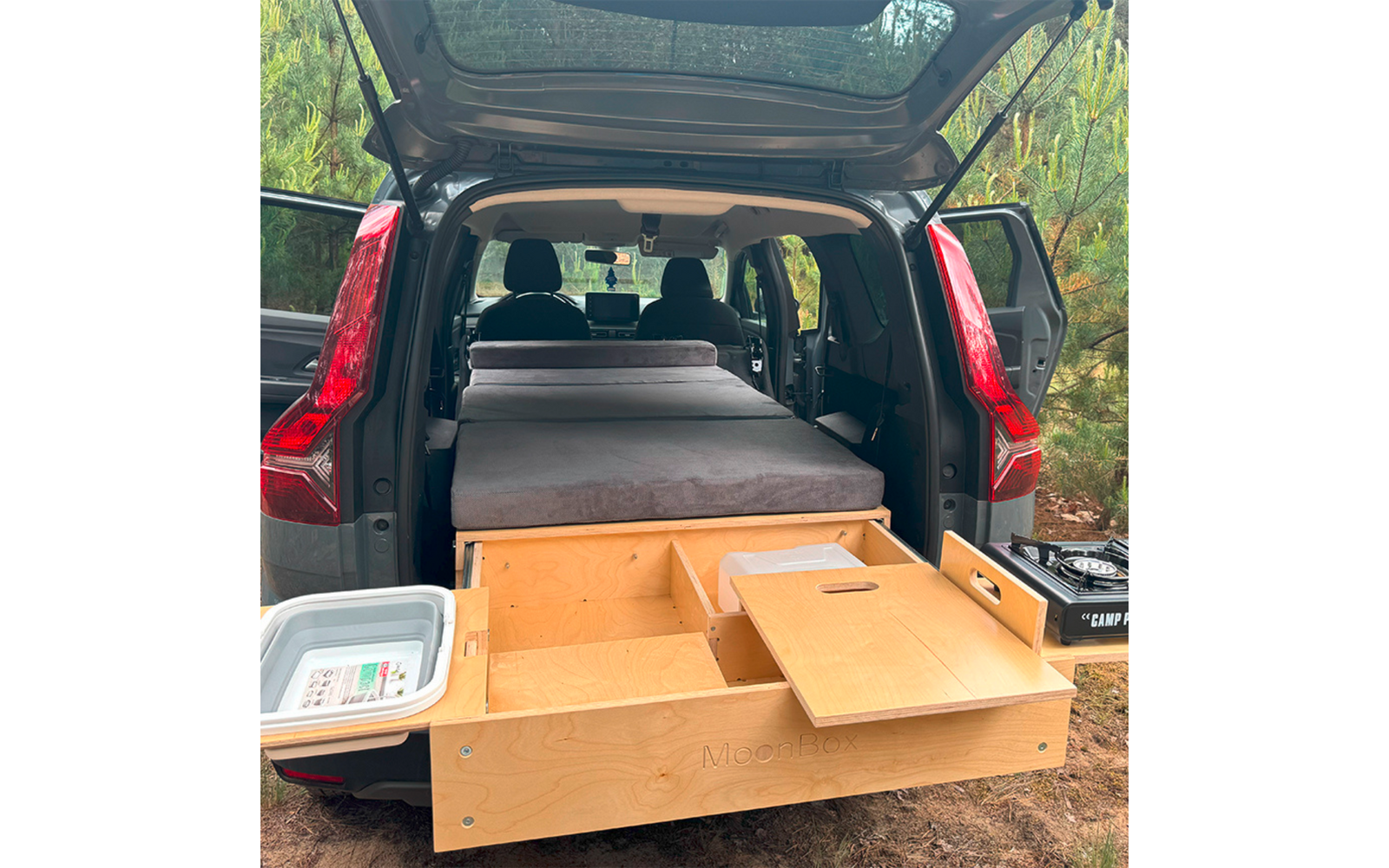 Offenes Heck eines SUVs mit grauer Matratze auf einer Plattform, herausziehbare Holzschublade mit Fächern, Plastikwanne links, Campingkocher rechts, Nadelbäume im Hintergrund
